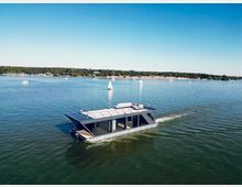 Ein modernes Hausboot mit einer großen Terrasse fährt über einen ruhigen See, umgeben von kleineren Segelbooten und einem Ufer mit Bäumen und Anlegestellen im Hintergrund.
