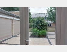 Blick durch eine Glastür auf einen gepflasterten Innenhof mit Beeten, die Gras und Büsche enthalten. Im Hintergrund sind Gebäudefassaden aus hellen Klinkersteinen mit geschlossenen Rollläden sowie ein flaches Blechdach sichtbar.