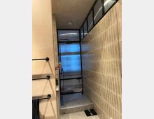 Moderne, beige geflieste Dusche mit Glas- und Metallabtrennung in einem Badezimmer. Der Boden besteht aus terrazzoartigem Material, mit einer ebenerdigen Duschkabine und einer großen, schwarzen Regendusche.