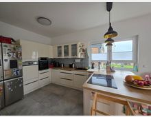 Moderne, helle Wohnküche in L-Form mit cremefarbenen Schränken, grauer Fliesenrückwand, eingebautem Backofen und großem Kühlschrank. Eine Arbeitsinsel mit Kochfeld steht vor einem breiten Fenster mit Plissees; darüber hängen schwarze Pendelleuchten.