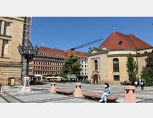 Ein städtischer Platz mit Pflastersteinen, umgeben von historischen Gebäuden, darunter ein gelbliches Gebäude mit roten Ziegeldächern und Rundbogenfenstern sowie ein mehrstöckiges Gebäude mit roten Fassaden. Im Vordergrund stehen eine Straßenlaterne, Steinbänke und eine Person sitzt auf einer Bank.