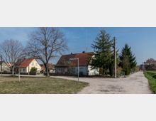 Ländliche Wohngegend mit Einfamilienhäusern, die rote und gelbe Dächer haben, umgeben von Bäumen und kleinem Vorgärten. Eine unbefestigte Straße führt an den Häusern entlang, und ein Straßenschild mit der Aufschrift „Heinrich-Heine-Straße“ ist zu sehen.