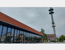 Moderne Feuerwehrstation mit großflächiger Glasfassade und einer roten Ziegeldachverkleidung. Im Hintergrund befindet sich ein hoher Kommunikationsmast sowie ein Backsteingebäude, umgeben von vereinzelten Bäumen und parkenden Autos.