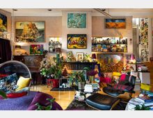 Wohnzimmer mit gemütlicher und bunter Gestaltung, gefüllt mit vielen Büchern, Pflanzen und Kunstwerken an den Wänden. Der Raum hat hölzernen Boden, eine Sitzecke, einen hängenden Acrylsessel, ein Klavier und zahlreiche Dekorationsgegenstände.