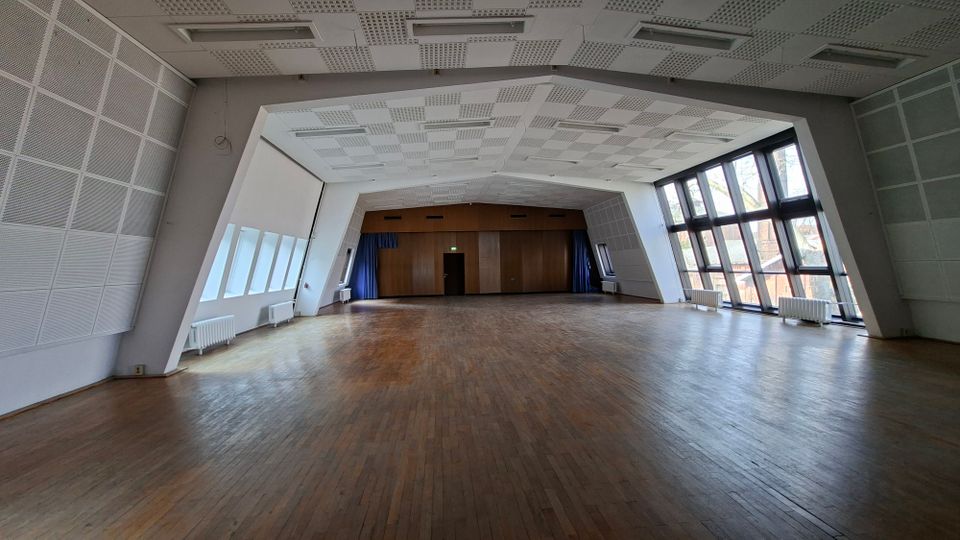 Großer, leerer Veranstaltungssaal mit Holzboden und hoher, schallgedämmter Decke mit eingelassenen Leuchten. Rechts sorgt eine hohe Fensterfront für Tageslicht, hinten ist eine holzverkleidete Stirnwand mit Tür und seitlichen Vorhängen zu sehen.