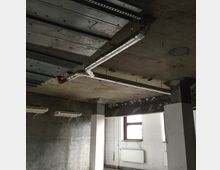 Rohbau-Interieur mit Betonwänden und -decken, sichtbaren Lüftungsrohren aus Metall und einer Reihe von Wandheizkörpern unter einem Fenster. Die Decke zeigt freiliegende Träger und Rohre, die einen unvollendeten Baustatus andeuten.