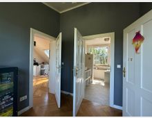 Ein heller Innenraum mit Blick auf zwei offene Türen, die jeweils in unterschiedliche Räume führen: links in ein Arbeitszimmer mit Dachfenster und rechts in eine Küche mit großen Fenstern. Im Vordergrund befindet sich ein Schrank mit Gesellschaftsspielen, und der Boden ist aus Holzparkett.