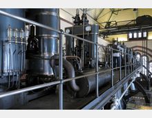 Innenraum einer historischen Industriehalle mit großen Dampfmaschinen und dunkelblauen Rohren, umgeben von metallischen Geländern. Im Hintergrund steht eine Person in Arbeitskleidung neben den Maschinen, während Tageslicht durch hohe Rundbogenfenster fällt.