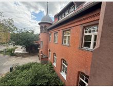 Außenansicht eines Backsteingebäudes mit zahlreichen rechteckigen Fenstern und einem Turm mit spitzer Kuppel. Im Vordergrund sind Büsche und ein Gehweg zu sehen, im Hintergrund Bäume und eine Straße bei bewölktem Himmel.