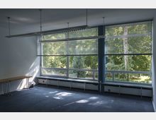 Ein leerer, moderner Raum mit großen Fenstern, durch die Bäume und eine grüne Parklandschaft sichtbar sind. Im Inneren befinden sich eine blaue Säule, hängende Leuchten sowie ein Teppichboden; einige zusammenklappbare Tische stehen an der Wand.