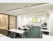 Seminarraum mit weißen Wänden, mehreren Tischen und Stühlen in Reihen angeordnet. Im Hintergrund ein großer Bildschirm mit der Beschriftung "Seminarraum (25 Personen)" sowie ein Flipchart; links ein abgetrennter Bereich mit einer Glaswand und bunten Plakaten.