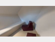 Minimalistisch gestalteter Innenraum mit schrägen weißen Wänden und zwei dunkelroten, geometrisch geformten Sitzmöbeln auf einem hellen Boden. Die Wände und Möbel schaffen eine abstrakte, moderne Atmosphäre.