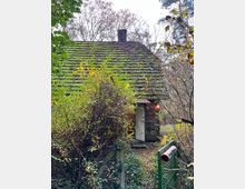 Kleines, von dichtem Gebüsch umgebenes Haus mit einem Moos bewachsenen Schrägdach, einem Schornstein und einer offenen Tür. Im Vordergrund ein grünes Gartentor mit einem schmalen Weg, der zum Gebäude führt.