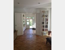 Helle Wohnraumansicht mit Parkettboden, geöffneten weißen Flügeltüren und einem Blick durch den Eingangsbereich auf eine grüne Hecke. Rechts steht ein weißes Regal mit Büchern und Dekorationen, links ein Sofa an der Wand.