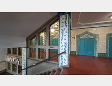 Innenansicht eines Treppenhauses mit breiten Fenstern in geometrischen Sprossenmustern und einem Treppengeländer entlang der Stufen. An den Wänden und Säulen sind helle Fliesen mit blau-weißen Ornamenten, gegenüber stehen türkisfarbene Doppeltüren mit dekorativ gerahmtem Türbogen.