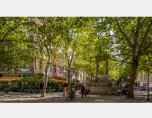 Ein kleiner Platz in einer urbanen Umgebung, gesäumt von dichten Bäumen, die Schatten spenden. Im Zentrum steht ein steinerner Brunnen mit Skulpturen, umgeben von Sitzbänken; im Hintergrund befinden sich ein Restaurant mit gelber Markise und Gebäude mit Wohn- und Geschäftseinheiten.