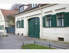 Wohnhaus in traditioneller Architektur mit hellen verputzten Wänden und grün gestrichenen Fensterläden. Vor dem Gebäude befindet sich ein gepflasterter Gehweg, und im Hintergrund sind weitere Häuser mit roten Ziegeldächern sowie ein Baum sichtbar.