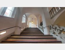 Breite Treppe in einem historischen Gebäude mit kunstvoll verzierten Geländern, einem hohen, gewölbten Deckenbereich und großen Fenstern auf der linken Seite, die natürliches Licht einfallen lassen. Eine hängende Laterne und eine Pflanze im oberen Bereich setzen Akzente.