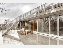 Moderne Terrasse mit glatter Betonoberfläche und großen Glasfronten, die in ein Gebäude führen. Links führt eine freistehende Metalltreppe nach oben, und auf der Terrasse stehen minimalistische Holzstühle. Hintergrund zeigt kahle Bäume und eine bewölkte Atmosphäre.
