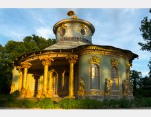 Pavillon im Rokokostil mit runder Grundform, verziert durch vergoldete Statuen, Säulen in Palmform und dekorative Fenster; umgeben von üppiger Vegetation und beleuchtet vom Abendlicht.