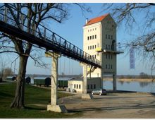 Industriegebäude mit hellem Putz und rotem Satteldach, durch das eine lange Metallbrücke mit Geländer führt, gestützt von Betonpfeilern. Im Hintergrund befinden sich eine Wiese, ein ruhiger Fluss und Güterwaggons.
