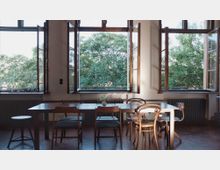 Helles Zimmer mit großen, offenen Holzrahmenfenstern, die den Blick auf üppige grüne Bäume freigeben. Im Vordergrund steht ein Esstisch mit unterschiedlichen Stühlen, dahinter befinden sich Heizkörper an der Wand.