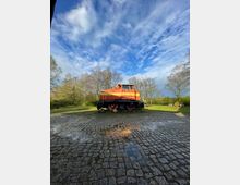 Eine rote Lokomotive steht auf einem gepflasterten Platz, umgeben von Bäumen und grünem Rasen unter einem teilweise bewölkten blauen Himmel. Auf dem Pflaster ist eine Wasserpfütze, in der sich die Lokomotive spiegelt.