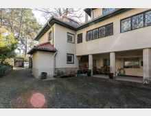 Zweistöckiges Wohngebäude mit heller Fassade, roten Ziegeldächern und Gitterfenstern. Vor dem Haus befinden sich ein gepflasterter Hof, Terrassenmöbel und umgebende Bäume sowie ein Holztor im Hintergrund.