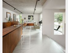 Open-plan modern living and dining area with a long wood cabinet and dining table, white walls, and a raw concrete ceiling with track lighting. Large floor-to-ceiling windows bring in garden views, with a sitting area, a grand piano, and modern chairs arranged throughout.
