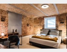 Schlafzimmer in einem historischen Gebäude mit freigelegten Backsteinwänden und einem gewölbten Ziegeldeckenbogen. Der Raum enthält ein modernes Doppelbett mit gelber Decke, einen Schreibtisch aus Holz und Metall sowie ein Fenster, das Tageslicht hineinlässt.