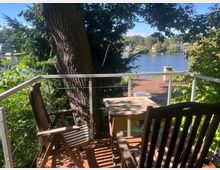 Holzterrasse mit zwei Stühlen und einem Tisch, umgeben von dichten Bäumen und Blick auf einen See im Hintergrund; im Vordergrund steht ein großer Baum, und im Seeuferbereich sind Häuser mit roten Dächern erkennbar.