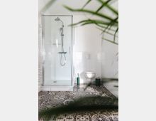 Modernes Badezimmer mit gläserner Duschkabine, hochgelegter Toilette und dekorativen Fliesen in Schwarz-Weiß-Mustern. Im Vordergrund sind unscharfe Pflanzen sichtbar, die eine natürliche Atmosphäre hinzufügen.