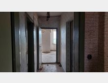 Ein verlassener Flur mit abgenutzten Wänden und Decke, die teilweise Farbe oder Tapete verloren haben. Am Ende des Flurs führt eine offene Tür in einen Raum mit einem Fenster, das Tageslicht hereinlässt und einen Blick nach draußen zeigt.