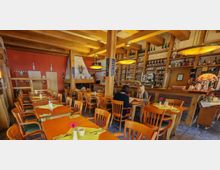 Ein gemütliches Restaurant mit Holzmöbeln und warmem Beleuchtungskonzept. Es gibt mehrere gedeckte Tische mit gelben Servietten und Blumen, eine Theke im Hintergrund mit Regalen voller Gläser und Flaschen sowie eine Ziegelwand und dekorative Elemente.