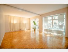 Heller Raum mit Parkettboden, großen Fensterfronten, die viel natürliches Licht hereinlassen, und cremefarbenen Vorhängen an einer Wand. Im Raum stehen vereinzelte Gegenstände wie eine Pflanze in einem Topf und zwei Stehlampen.