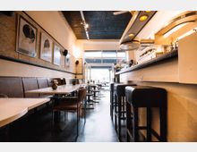 Ein modernes Café mit einer langen Bar auf der rechten Seite und Sitzbereichen auf der linken Seite, bestehend aus Tischen, Stühlen und einer gepolsterten Bank. An der Wand hängen gerahmte Schwarz-Weiß-Fotografien, und im Hintergrund führt eine gläserne Eingangstür nach draußen.