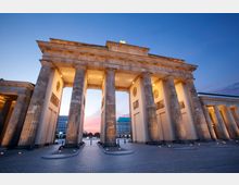 Das Bild zeigt das Brandenburger Tor in Berlin bei Dämmerung. Die beleuchteten Säulen und Verzierungen des historischen Bauwerks heben sich vor einem blauen Himmel und einem rosa Lichtstreifen im Hintergrund ab.