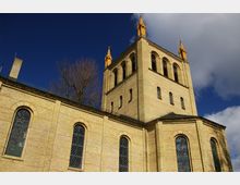 Backsteinkirche mit romanischen Rundbogenfenstern und einem quadratischen Turm, dessen Spitze mit auffälligen gelblichen Zierelementen verziert ist, vor einem blauen Himmel mit weißen Wolken. Ein Baum ohne Blätter ragt hinter dem Gebäude hervor.
