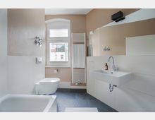 Helles Badezimmer mit einer Wand in Beige- und Weißtönen, ausgestattet mit einer Badewanne, einem Hänge-WC, einem Waschbecken mit Spiegel sowie einem Handtuchheizkörper. Das Fenster mit Blick auf ein Nachbargebäude sorgt für zusätzliche Beleuchtung, während dunkelgraue Bodenfliesen Akzente setzen.