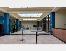 Heller Innenraum eines modernen Gebäudes mit einer Glasdachkonstruktion, runden Stehtischen und blauen Säulen. Rechts im Bild befinden sich raumhohe Fenster und Holzwandverkleidungen, links ist eine Treppe zu sehen.