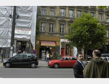 Straßenszene mit einem historischen Gebäude, dessen Fassade teils renoviert und teils von Bauplanen bedeckt ist. Im Erdgeschoss befinden sich Geschäfte, darunter ein Laden mit der Aufschrift „Koof im Kiez!“, davor parkende Autos und Gehwegpassanten.