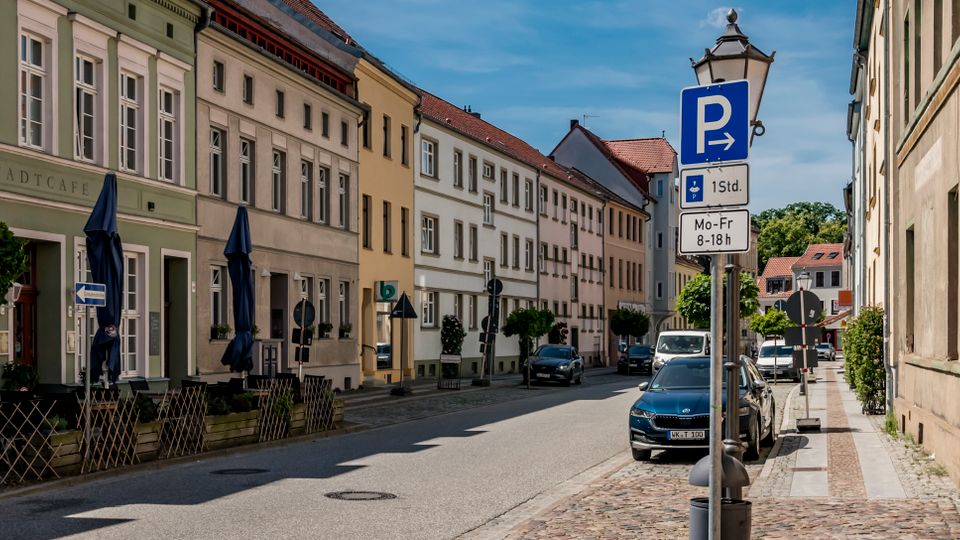 Straßenzug in einer historischen Altstadt mit mehrgeschossigen, pastellfarbenen Fassaden und hohen Fenstern, teils mit Caféfront im Erdgeschoss. Am rechten Straßenrand stehen parkende Autos, ein Laternenmast mit Parkhinweisen sowie Gehwege mit Kopfsteinpflaster und kleinen Straßenbäumen.
