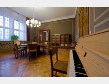 Ein stilvoll eingerichteter Raum mit Holzboden, einem Klavier im Vordergrund, einem runden Tisch mit Stühlen in der Mitte und einer Wand mit Bücherregalen. Große Fenster mit Vorhängen und ein Kronleuchter tragen zur klassischen Atmosphäre bei.