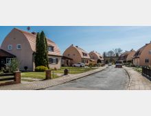 Straßenansicht eines Wohngebiets mit freistehenden Einfamilienhäusern im Bauhaus-Stil, die charakteristische abgerundete Dächer und helle Fassaden haben. Vor den Häusern befinden sich gepflegte Vorgärten, und die Straße ist mit Pflasterwegen und parkenden Autos gesäumt.