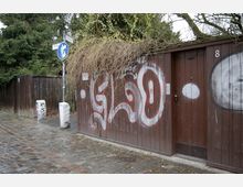 Ein gepflasterter Gehweg neben einem Holzzaun und einem Holztor, das mit roter und weißer Graffiti besprüht ist. Im Hintergrund sind Bäume und Schilder zu sehen, die eine verkehrsberuhigte Zone markieren.