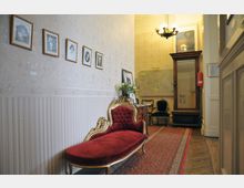 Ein eleganter Eingangsbereich mit einer roten, gepolsterten Chaiselongue mit goldenen Verzierungen, einer gemusterten Tapete, gerahmten Schwarz-Weiß-Fotografien an der Wand und einem antiken Holzspiegel. Im Hintergrund stehen ein Schreibtisch, Stühle und eine große Karte an der Wand, auf einem roten orientalischen Teppich.