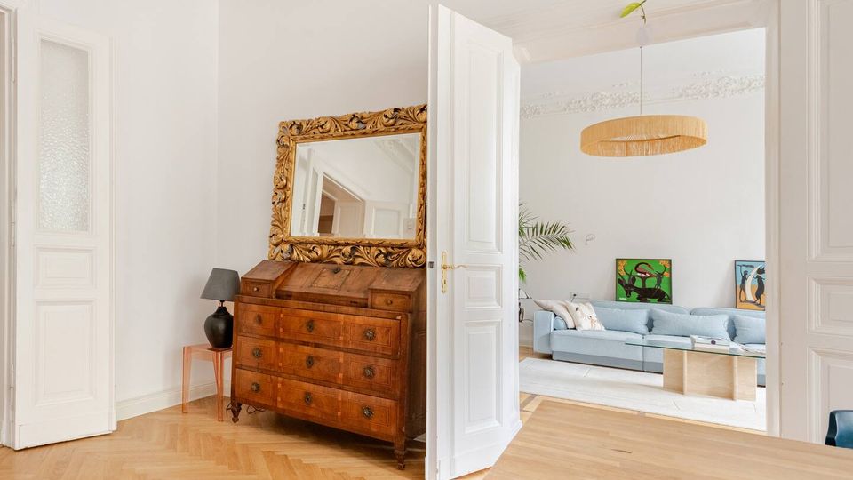 Heller Wohnungsflur mit Fischgrätparkett und weißen Flügeltüren, neben einer antiken Holzkommode mit großem, goldverziertem Spiegel. Durch die geöffnete Tür ist ein Wohnzimmer mit hellblauem Sofa, Glastisch und runder Deckenleuchte zu sehen.