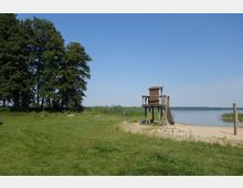 Grüne Wiese mit Bäumen am linken Bildrand, daneben ein Sandstrand mit einem hölzernen Kinderspielhaus mit Rutsche, das am Ufer eines ruhigen Sees steht. Im Hintergrund sind Schilf und eine bewaldete Uferlinie sichtbar.