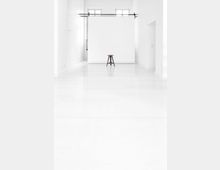 Ein minimalistisches, weißes Studio mit glattem Boden, einem einfachen Hocker in der Mitte und einer weißen, bodenlangen Hintergrundleinwand vor zwei hohen Fenstern.