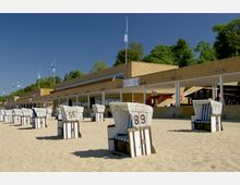 Ein Sandstrand mit mehreren weißen Strandkörben, im Hintergrund ein Gebäude aus gelbem Stein mit überdachten Arkaden, darüber wehen mehrere blau-weiße Fahnen. Dahinter sind grüne Bäume und ein klarer blauer Himmel zu sehen.